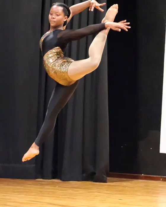 A dancer in a gold and black costume in a high leap with one leg extended back.
