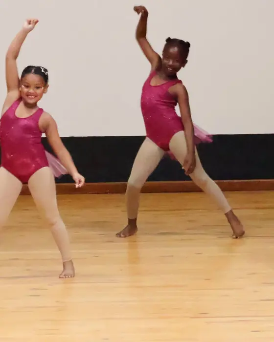 Young dancers in pink leotards with arms raised during a group routine.