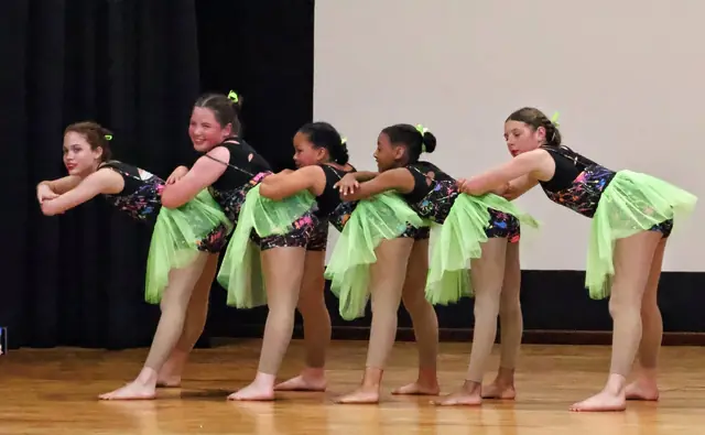 A line of dancers in green tutus leaning forward in a group routine.