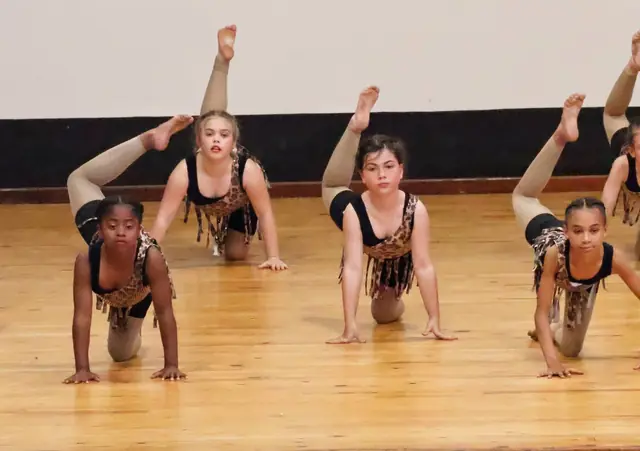 A group of dancers in leopard costumes kneel forward with one leg pointed high.