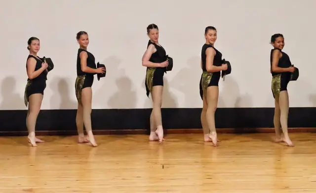 Five dancers in black and gold standing in a line in profile.