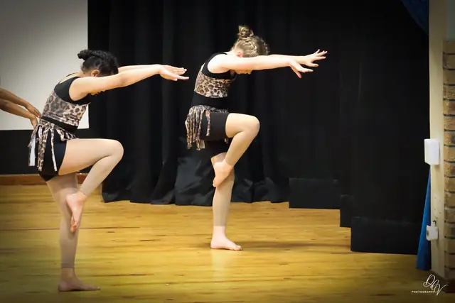 Two dancers in black and gold balancing on one leg with arms stretched forward.