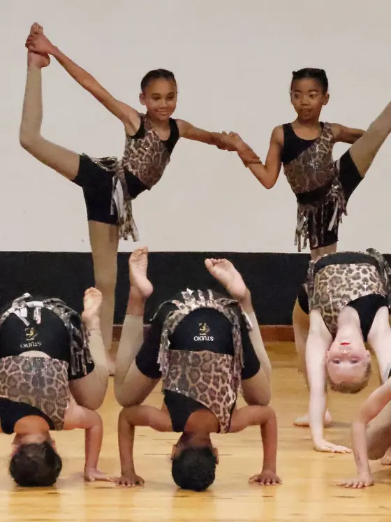 Dancers in leopard and gold costumes form an acrobatic pyramid with legs extended high.