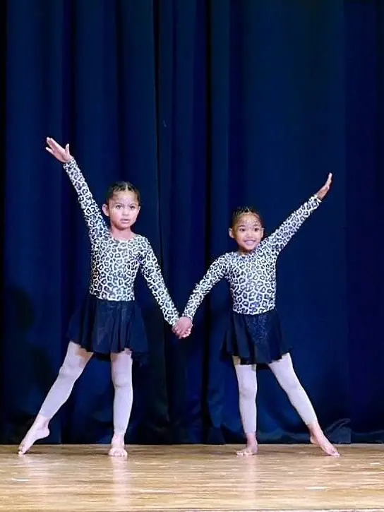 Two young dancers in matching leopard print outfits pose with arms raised on a blue lit stage.
