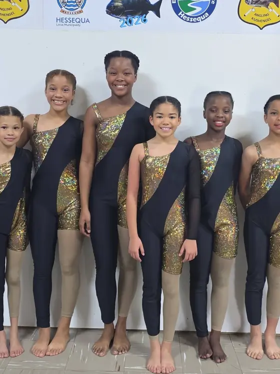 Six Studio E dancers in black and gold costumes stand in front of the Allegretto Eisteddfod sponsor board.