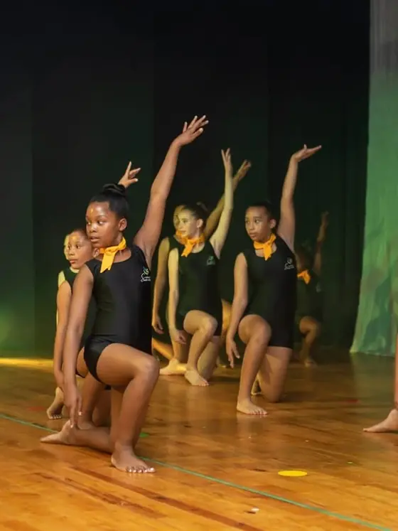 Dancers in black leotards and teal belts perform on a stage with green and gold lights.