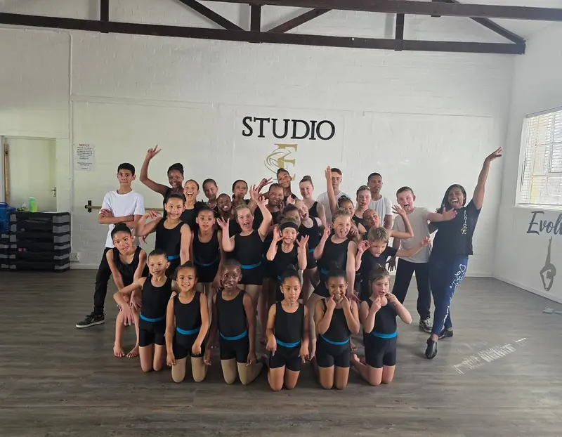 The Studio E dancers posing together in the studio, some with arms raised, in front of the gold logo.