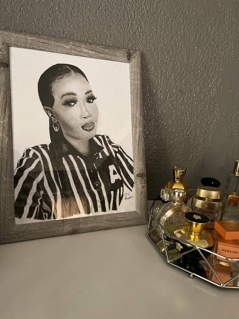 A finished graphite portrait of a woman in a striped shirt, framed in grey wood on a dresser