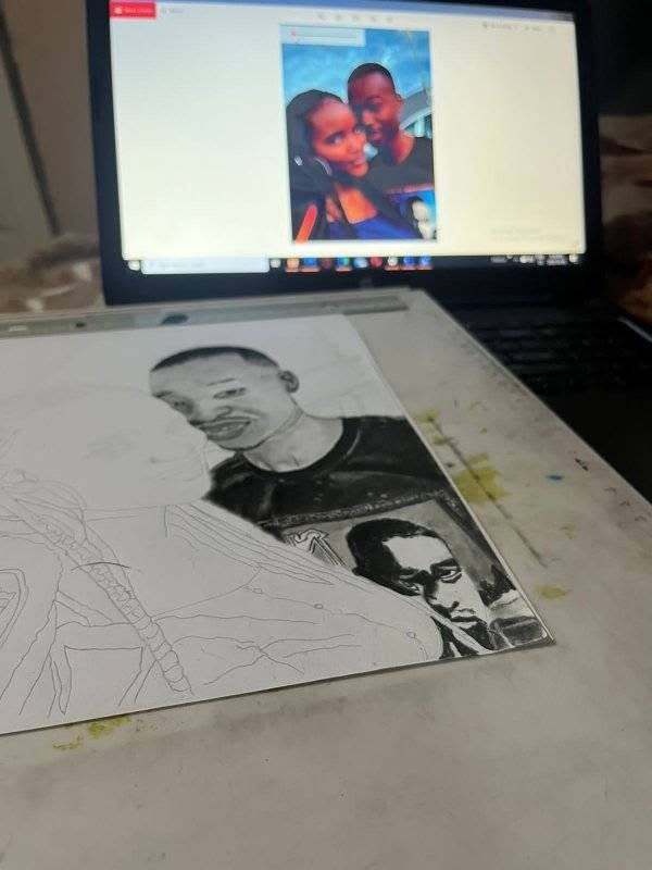 A graphite portrait half finished on the drawing desk, with the reference photo open on a laptop behind it